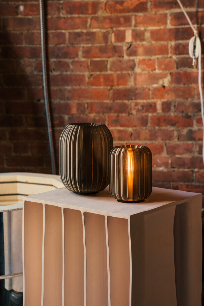 Two ribbed glass vases, crafted with Wooj Design elegance, stand on a textured white pedestal against a red brick wall—one larger and dark, the other smaller and lighted