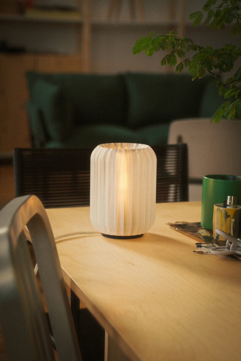 A white, pleated lampshade from Wooj Design graces a lit lamp on a wooden table, accompanied by green and white mugs and a book. In the background, a black chair complements the setting alongside a green sofa