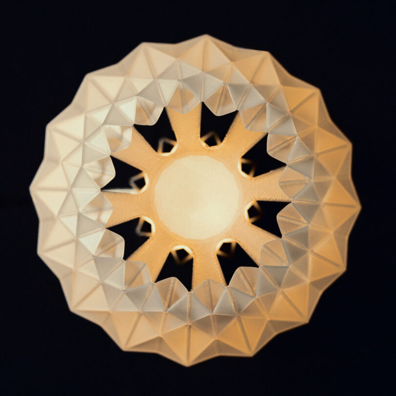 Close-up of a geometric, star-shaped lamp by Wooj Design, emitting warm light against a dark background. The lamp features a repetitive triangular pattern