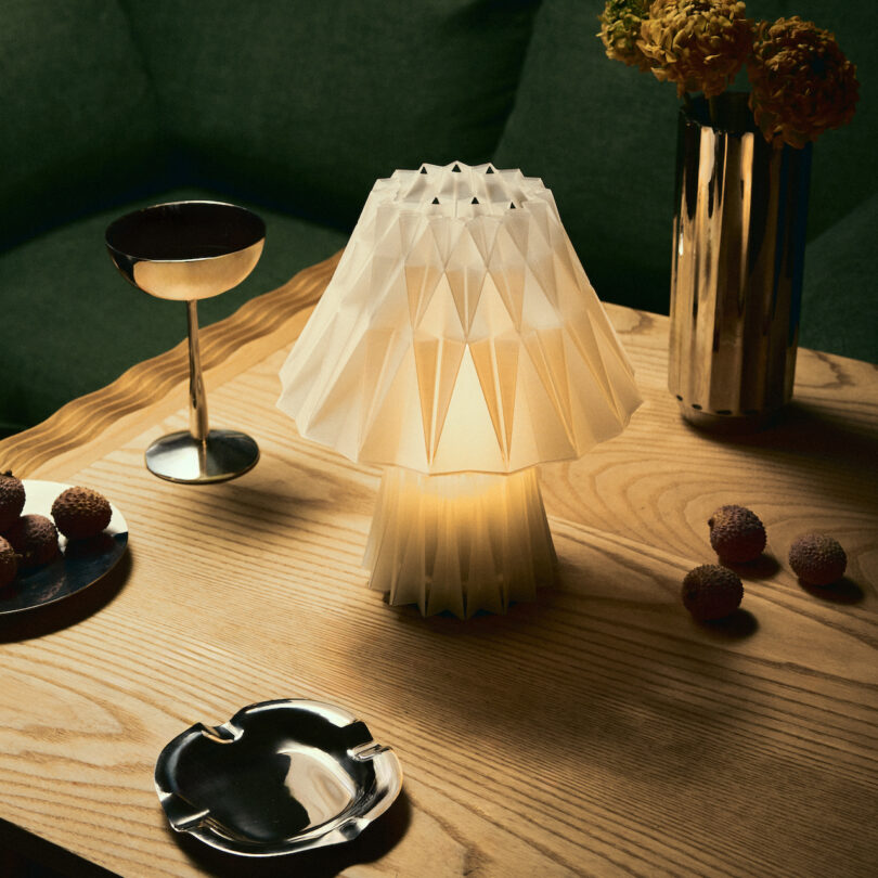 A geometric paper lamp by Wooj Design graces a wooden table, accompanied by a metallic drink, vibrant yellow flowers in a vase, and scattered fruit