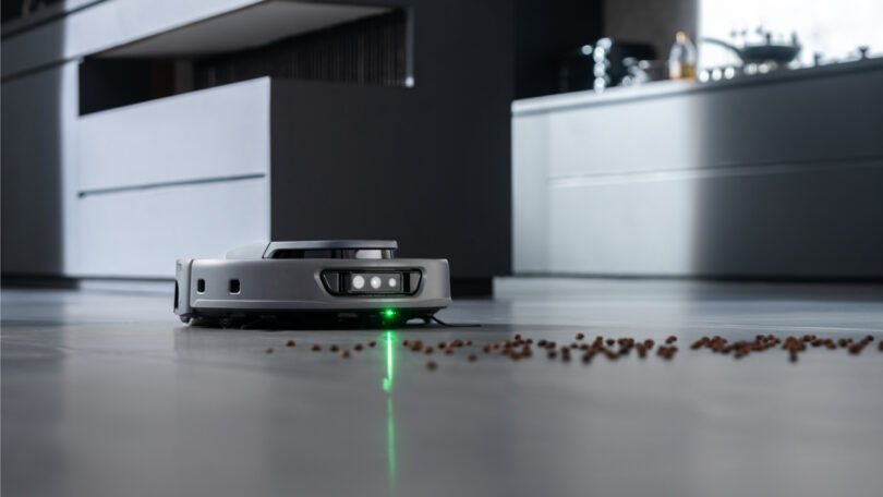 A robot vacuum cleans scattered debris off a tiled floor in a modern kitchen setting.
