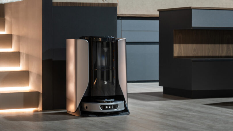 A robot vacuum cleaner docked at its charging station beside a set of illuminated stairs in a modern home interior.