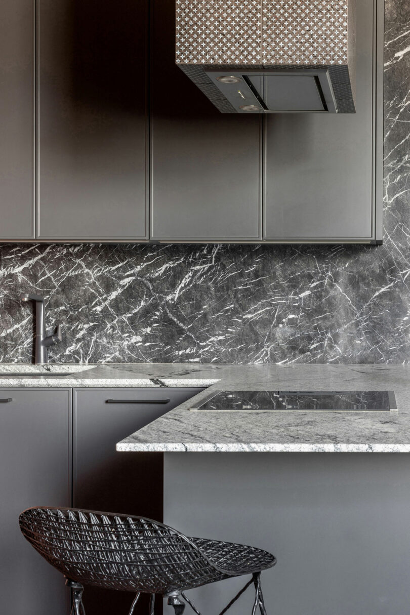 Modern kitchen with dark gray cabinets, a black marble backsplash, and a gray countertop. A black woven chair is placed near the counter, and an overhead range hood is visible.