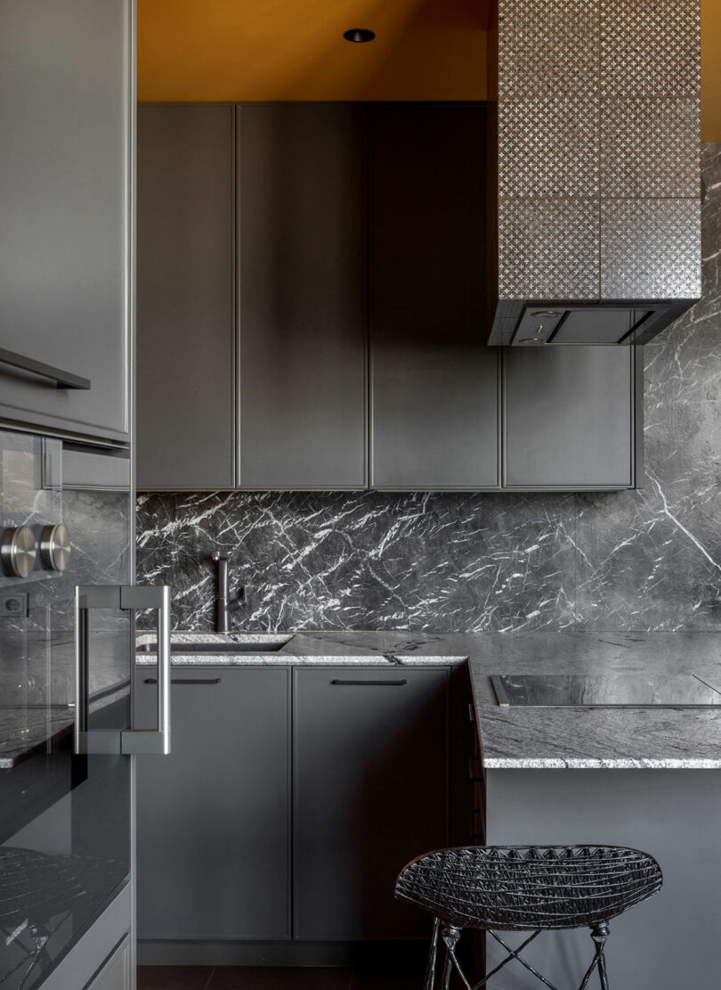 Modern kitchen with dark gray cabinets, marble backsplash, and countertops. Includes a stainless steel range hood, electric cooktop, and a black wireframe stool. Ceiling painted yellow.