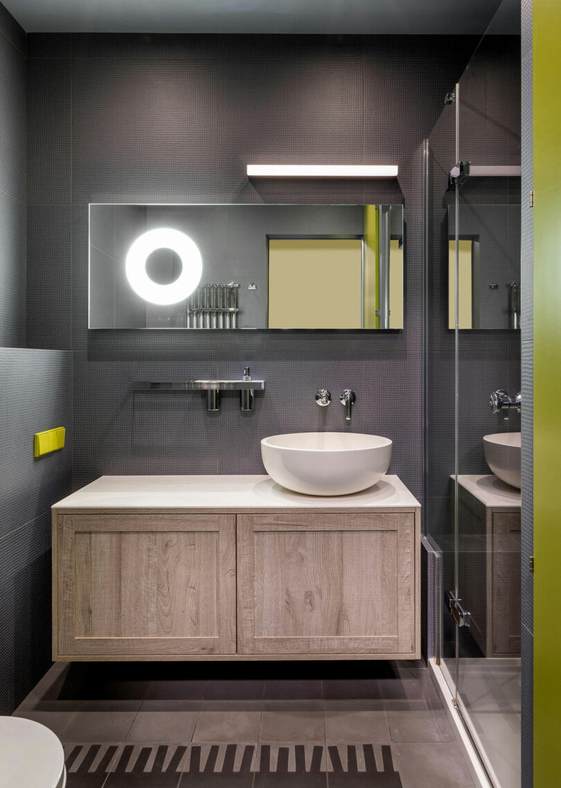 Modern bathroom with dark walls, round mirror with light, minimalist vanity, vessel sink, and glass shower enclosure.