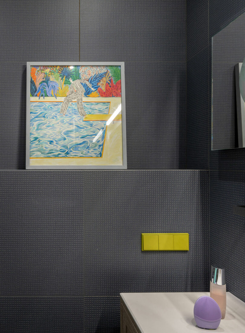 Framed abstract artwork above a bathroom sink with dark tiles and a small purple speaker.