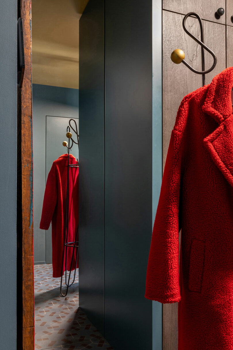 Red coat hanging on a wooden hook, reflected in a nearby mirror. Walls are painted dark green, with a patterned floor visible.