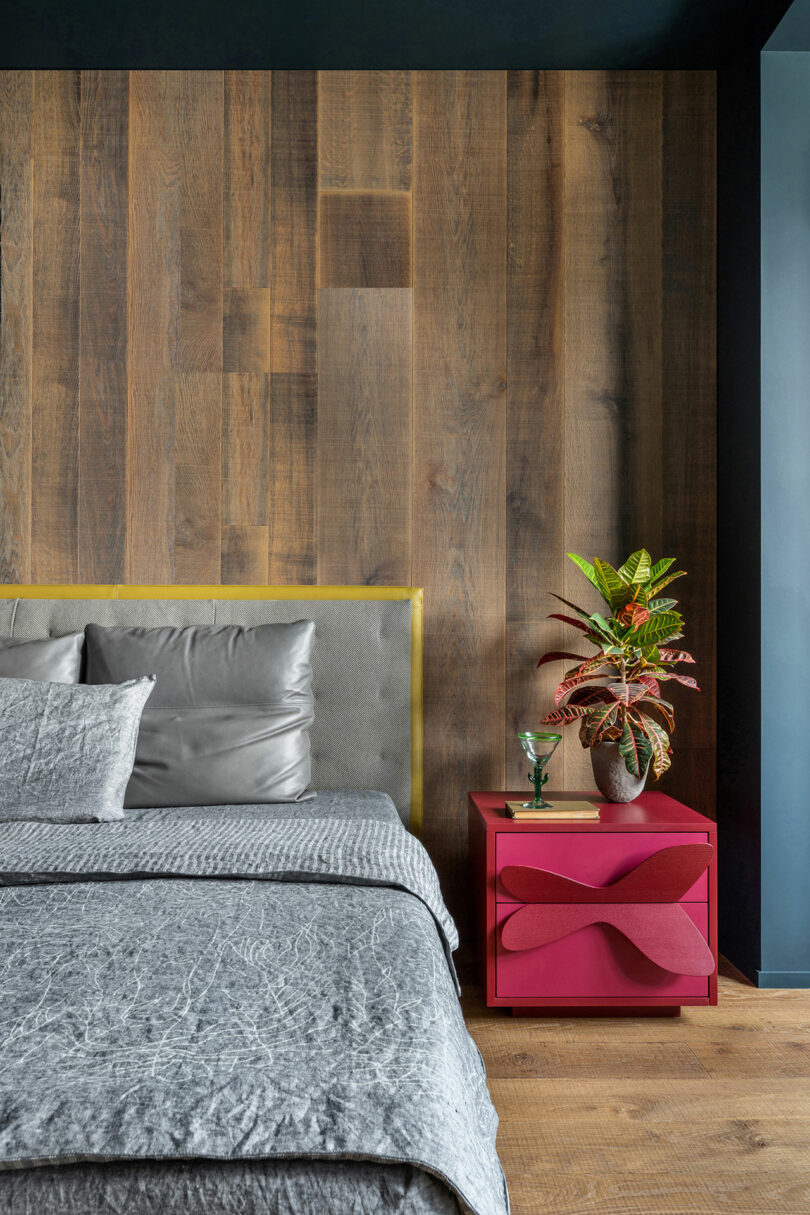 Modern bedroom with a gray upholstered bed against a wooden accent wall, gray bedding, and a pink nightstand holding a potted plant and a glass.