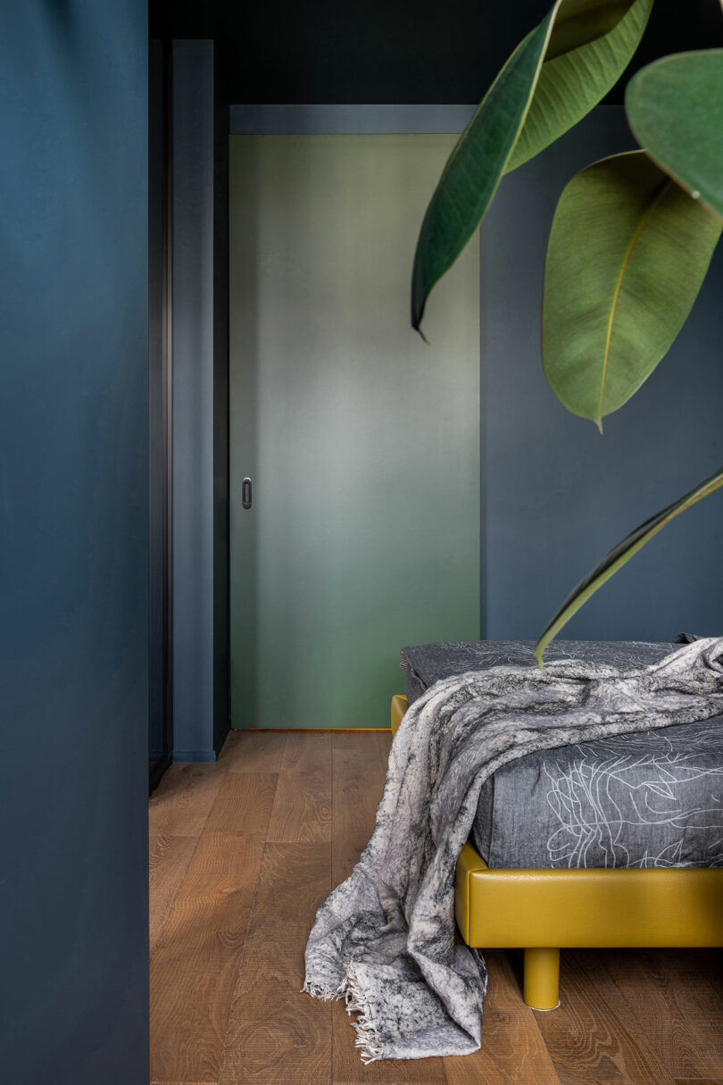 Modern bedroom with a muted green gradient sliding door, yellow bed base, and gray bedding. A large plant is in the foreground, and natural wood flooring is visible.