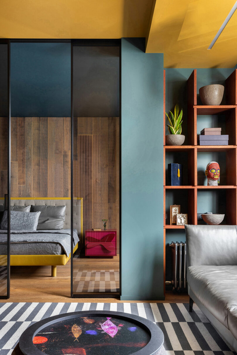 Modern living area with gray sofa, striped rug, and round black coffee table. View into bedroom with wood wall and yellow bed. Blue-green wall and wooden shelves with decor pieces visible.