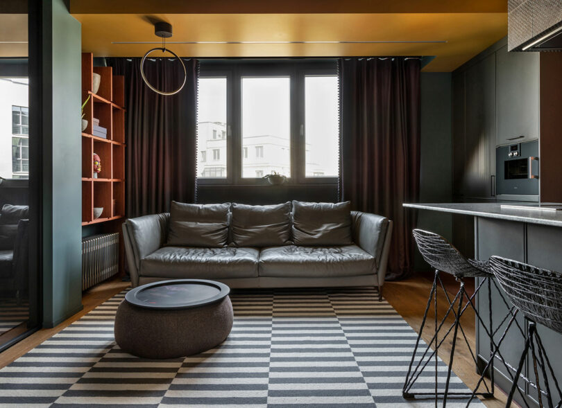 Modern living room with a gray sofa, round coffee table, striped rug, and bar stools. Dark curtains cover windows, and a wooden shelf is in the corner. Ceiling light fixture is overhead.