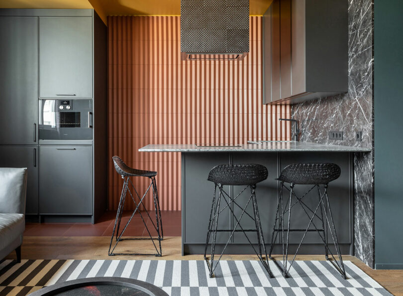 Modern kitchen with a marble countertop island, two black woven bar stools, textured orange backsplash, gray cabinets, and built-in appliances. A striped rug is on the wooden floor.