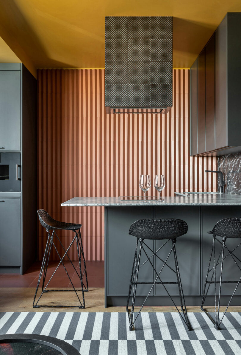 Modern kitchen with dark cabinetry, a marble countertop, two black bar stools, and a striped rug. Background features a textured, vertical pattern wall and a geometric range hood.