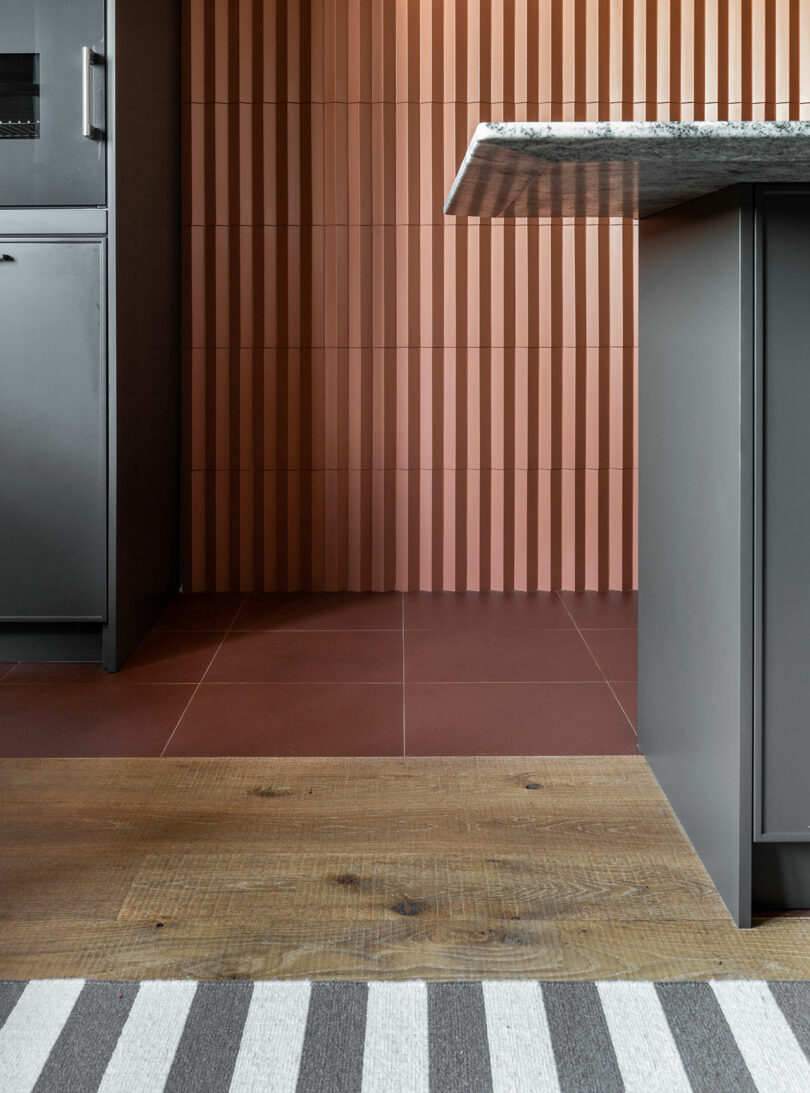 Modern kitchen interior with ribbed red wall tiles, dark cabinetry, and a granite countertop. The floor features wood and striped patterns.