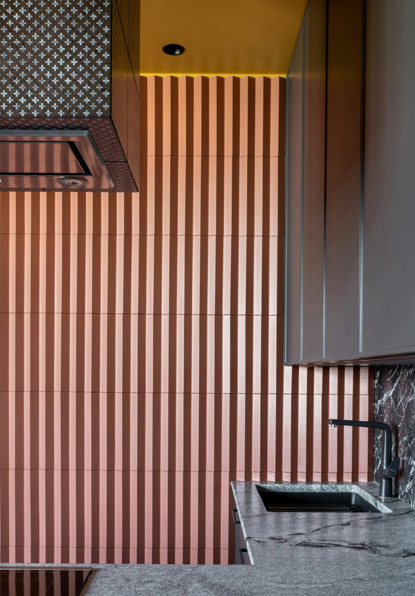 Kitchen interior with vertical orange striped wall tiles, gray countertops, a metal range hood, and a sink beneath gray cabinets.