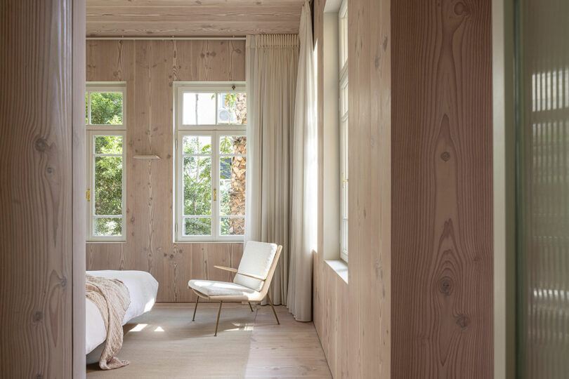 A cozy bedroom with wooden walls, featuring a bed, a chair, and large windows letting in natural light. Cream curtains and a blanket complete the serene and minimalist decor.