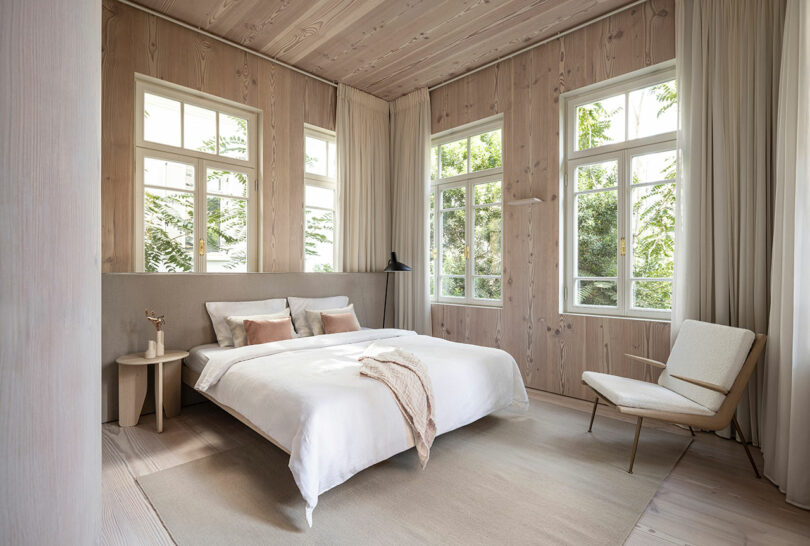 Minimalist bedroom with a large bed, neutral tones, and floor-to-ceiling windows showing greenery outside. A chair and side table complement the serene space.