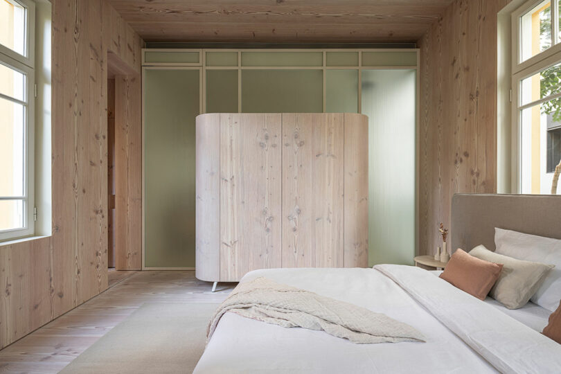 Minimalist bedroom with light wood paneling, a white bed, and a curved wooden wardrobe. Large windows provide natural light. Neutral tones dominate the decor.