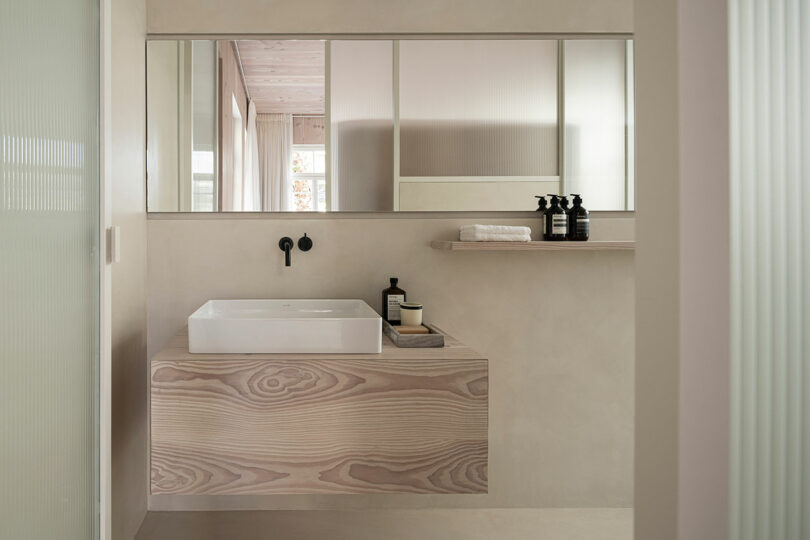 Minimalist bathroom with light wood vanity, rectangular sink, black faucet, large mirror, and a shelf with bottles and towels. Neutral color palette and frosted glass accents.