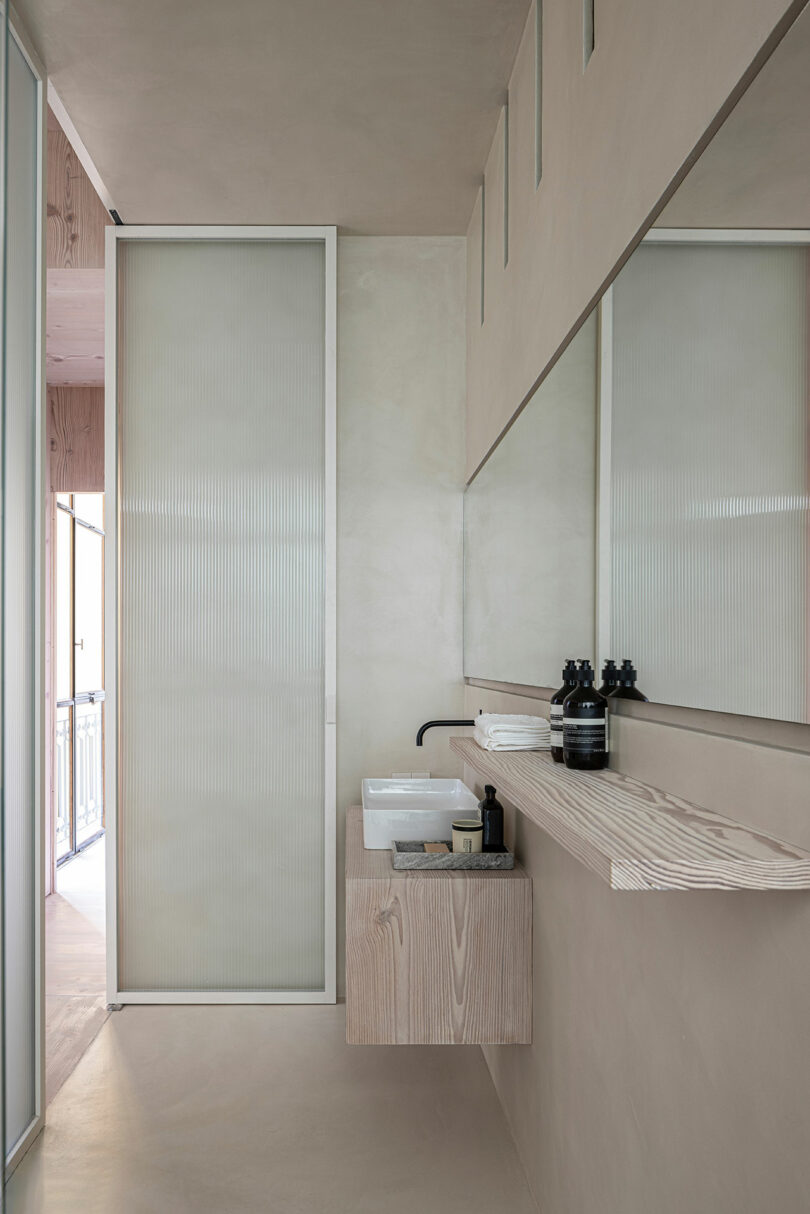 Minimalist bathroom with a rectangular basin, wall-mounted faucet, and wooden shelf holding toiletries. A frosted glass sliding door and large mirror are visible. Neutral color palette.