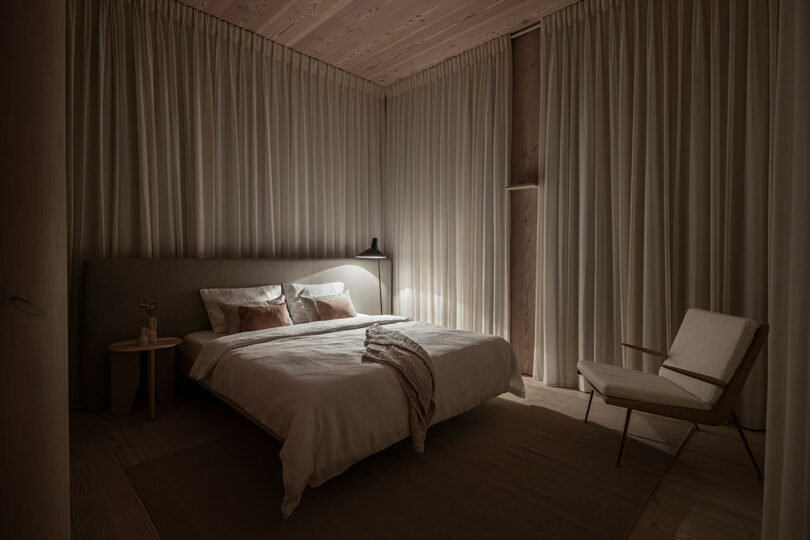 A dimly lit bedroom features a bed with white bedding, a side table with a lamp, and a cushioned chair. Beige curtains cover the walls, and the space has a minimalist design.