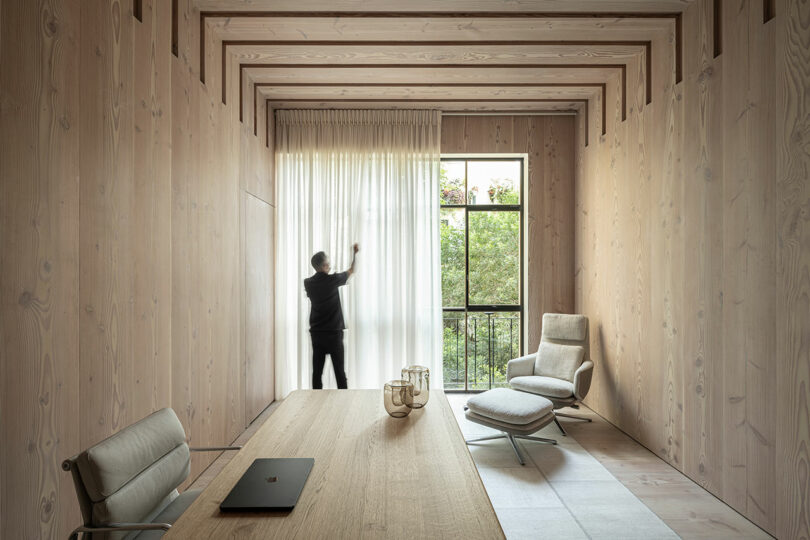 A person adjusts a curtain in a minimalist room with wood walls, a desk, two chairs, and a large window showing greenery outside.