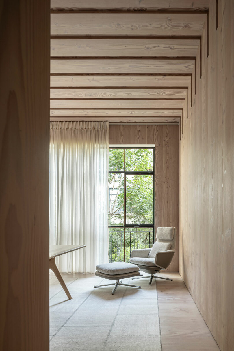 Minimalist room with wooden walls and ceiling beams, featuring a cushioned armchair, ottoman, and a window with sheer curtains overlooking greenery outside.