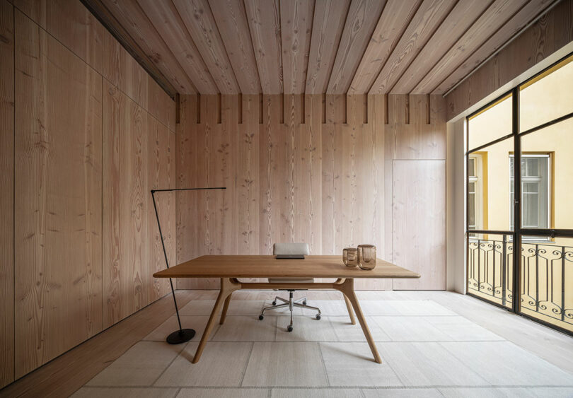 Minimalist office with a wooden desk, chair, and floor lamp. The room features wood-paneled walls and ceiling, a light-colored rug, and a large window with a balcony view.