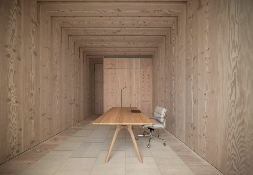 Minimalist wooden office space with a single desk, chair, and lamp. Walls, floor, and ceiling made of light wood with a textured pattern.