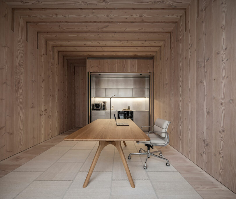 Minimalist wooden office room with a central table and white office chair. A wall unit with shelves and a coffee machine is in the background.