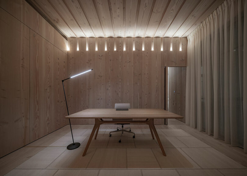 A minimalistic office with a wooden desk, swivel chair, floor lamp, and wall lighting. The room features wooden paneling, a carpeted floor, and sheer curtains.