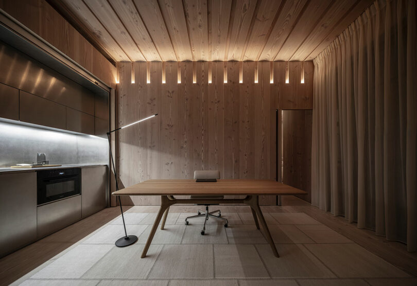 Minimalist office with a wooden ceiling and walls, modern wooden desk and chair, floor lamp, kitchenette on the left, and curtain on the right. Soft lighting from above.