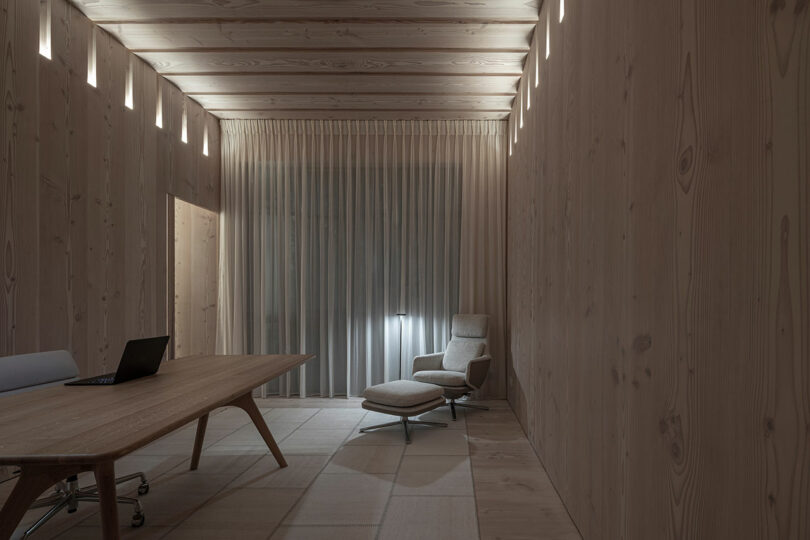 Minimalist wooden room with a long table, black laptop, and an armchair with an ottoman. Vertical windows near the ceiling provide soft lighting, and a curtain covers a large window.