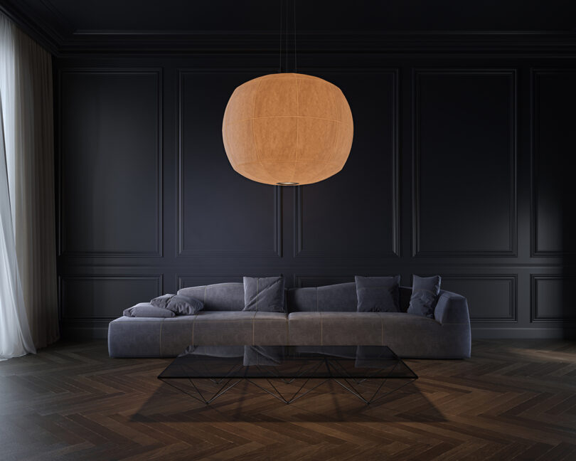 A dark-toned living room features a large, gray sectional sofa, a black geometric coffee table, and a warm, glowing spherical pendant light hanging above. The walls are elegantly paneled.