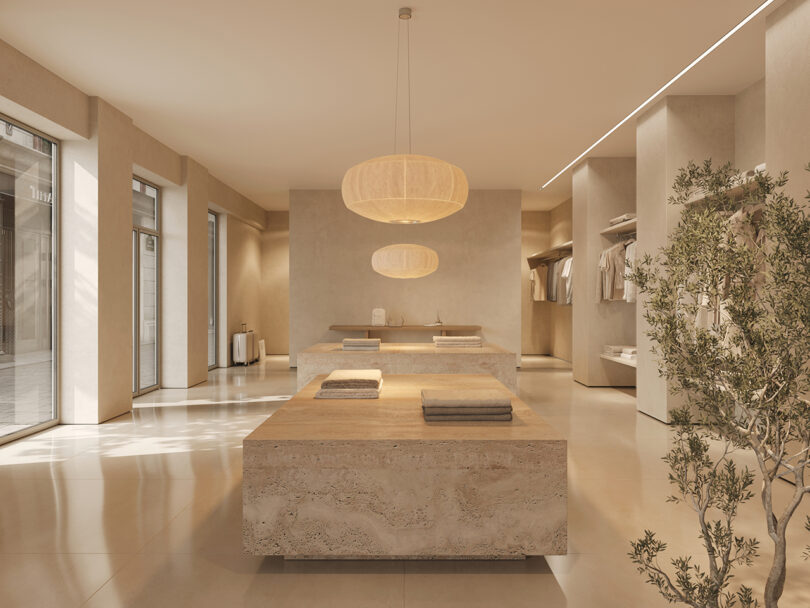 Minimalist clothing store with beige tones features wooden tables, hanging rounded lights, and neatly arranged clothes. Large windows allow natural light, and a potted plant adds greenery.