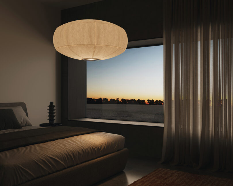 A bedroom with a large window view of a sunset. The room features a bed, a pendant lamp, sheer curtains, and minimal decor.