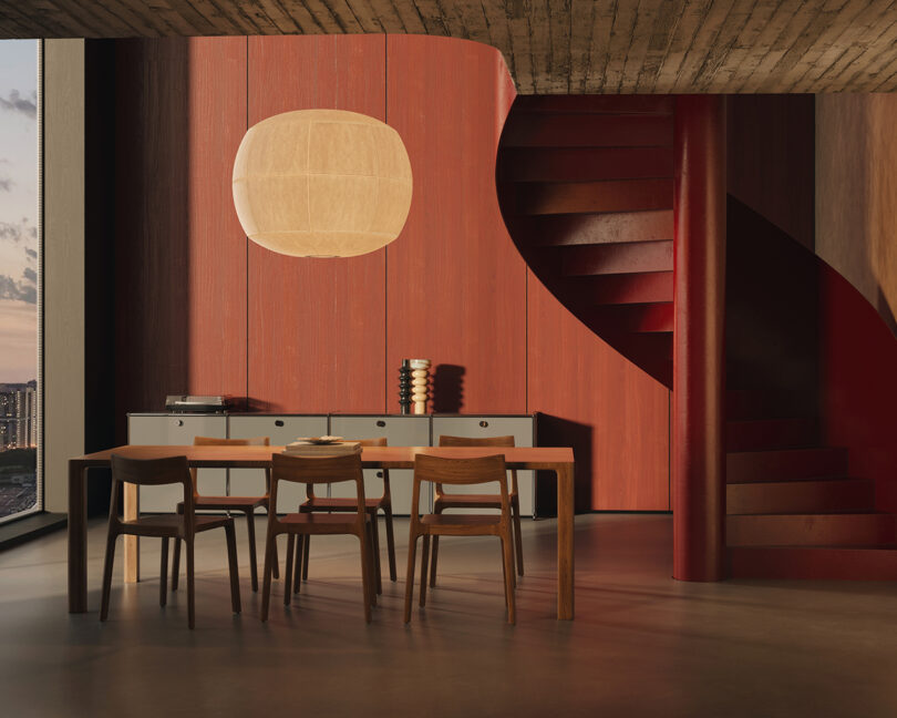 A dining area with a wooden table and four chairs, inside a room with red walls and a spiral staircase. A large round light fixture hangs above a sideboard. A cityscape is visible through the window.