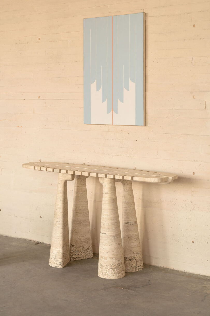 A modern, textured console table with unique conical legs stands against a plain wall. Above it hangs an abstract, symmetrical artwork in blue, light gray, and pink tones