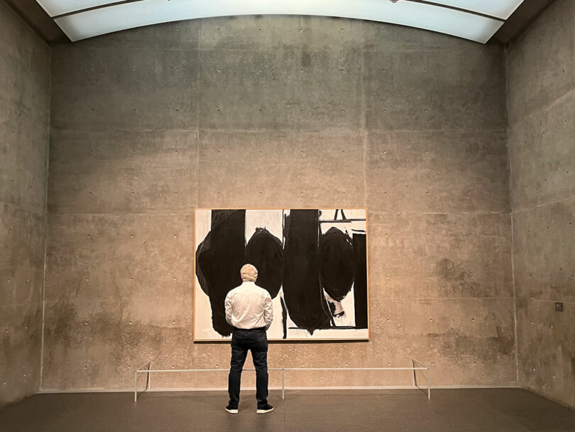 A person stands in front of a large abstract painting with black shapes in a concrete-walled gallery.