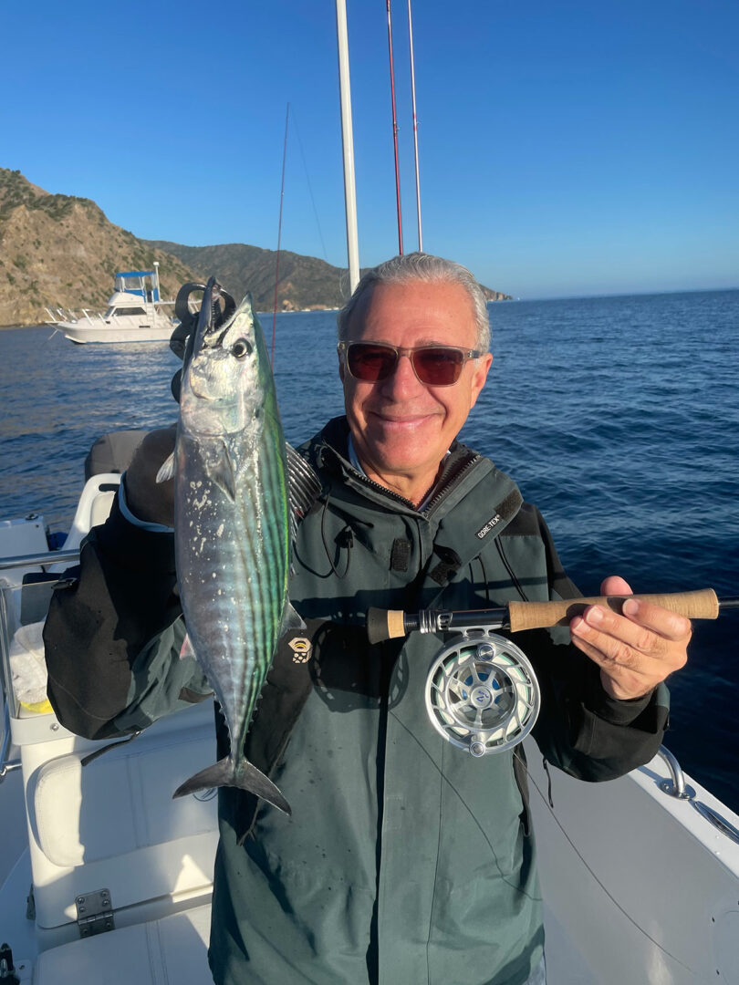 Man holding a fishing rod and a caught fish on a boat, with a mountainous landscape and other boats in the background.