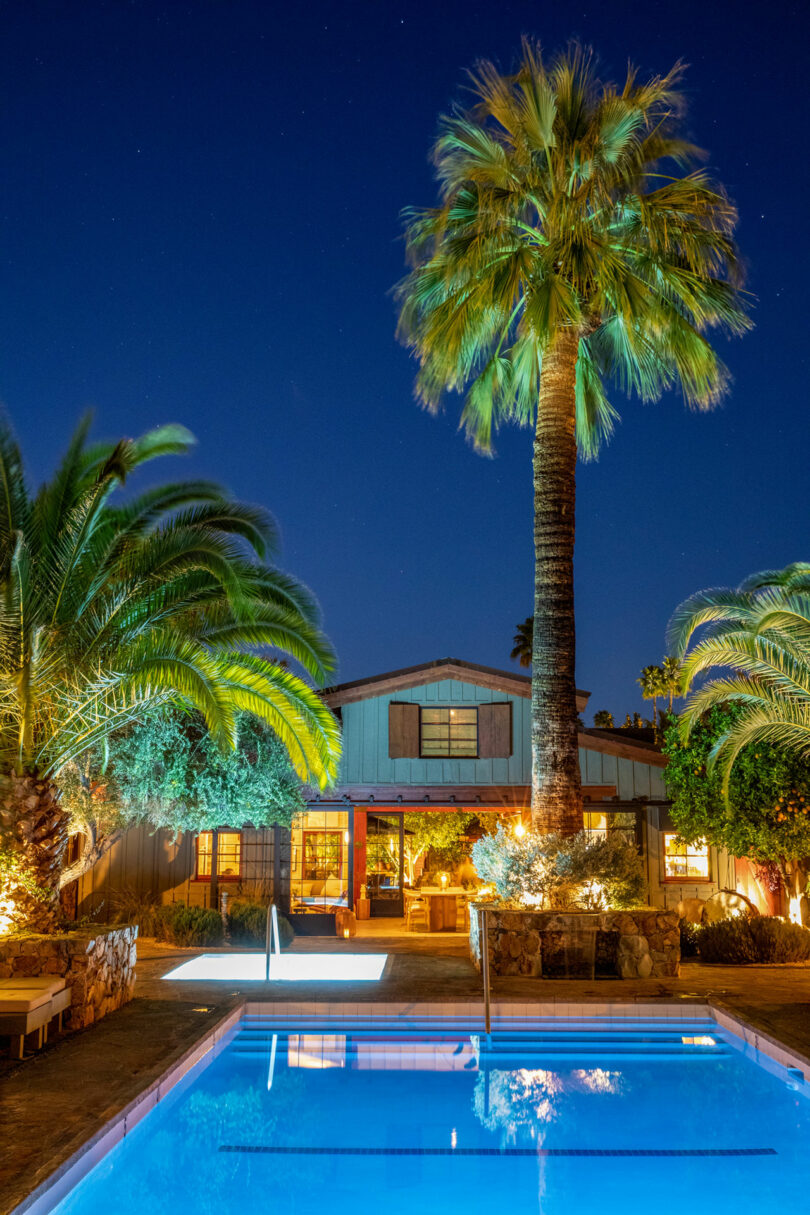 A house at night with illuminated windows and two palm trees. A swimming pool in the foreground reflects the lights and trees.