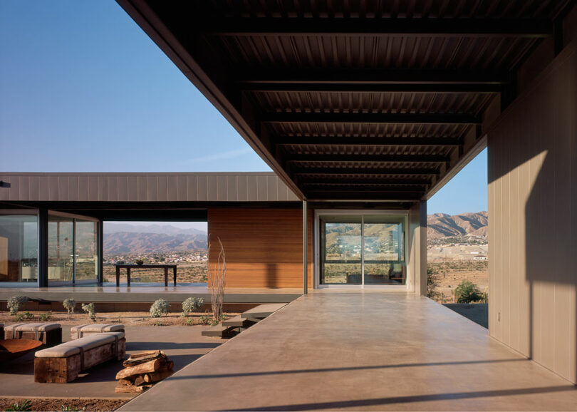 Modern architectural structure with open terrace and large windows overlooking a mountainous landscape under a clear blue sky. Minimalist design with concrete and metal elements.