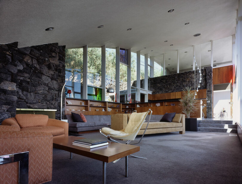 Modern living room with stone walls, large windows, and a mix of mid-century furniture, including sofas, a lounge chair, and a wooden coffee table. Natural light fills the space.