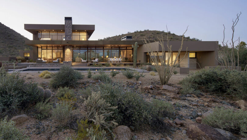 Modern house with large windows surrounded by desert landscape, including cacti and shrubs, set against a backdrop of hills under a clear sky.