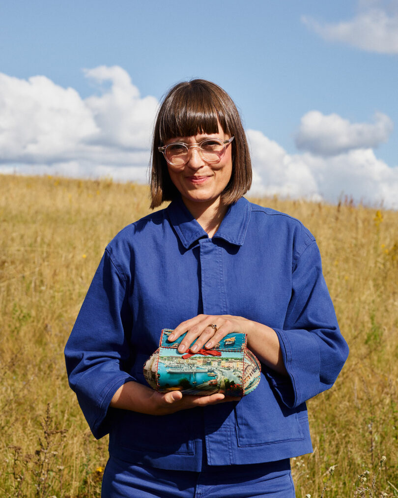 Stine Find Osther, clad in a blue jacket, stands in a field holding a small blue and green striped fabric object. A cloudy sky provides the backdrop.