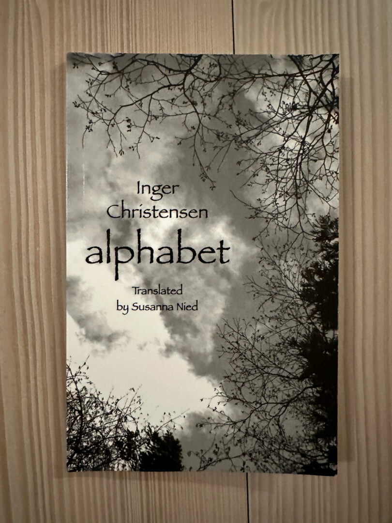 Cover of the book "alphabet" by Inger Christensen, translated by Susanna Nied, featuring a black and white sky and tree branches. The evocative artwork complements Stine Find Osther's interpretation, capturing the profound simplicity of nature's expanse.