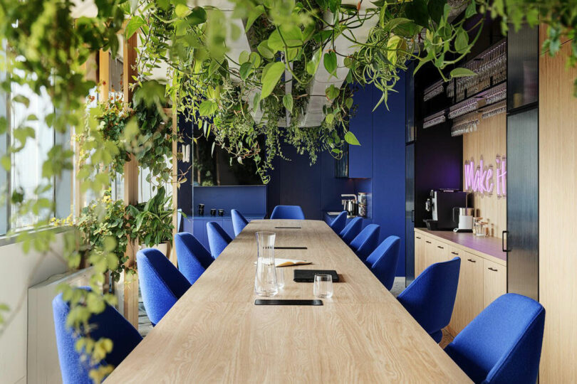 A long wooden conference table with blue chairs, surrounded by hanging green plants. Cabinets and a neon "Make It" sign are visible in the background.