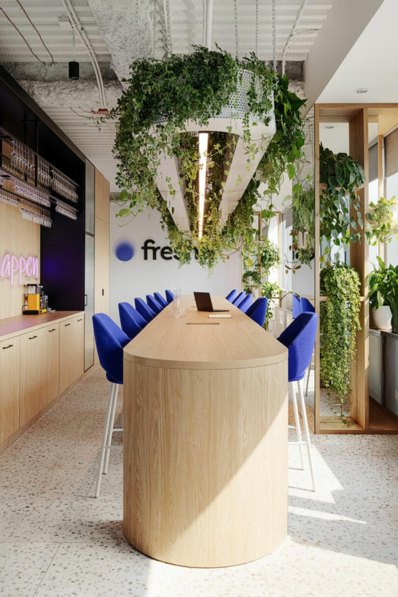 Modern office kitchen with wooden counters, blue bar stools, and hanging plants. A logo and neon sign are visible on the wall.