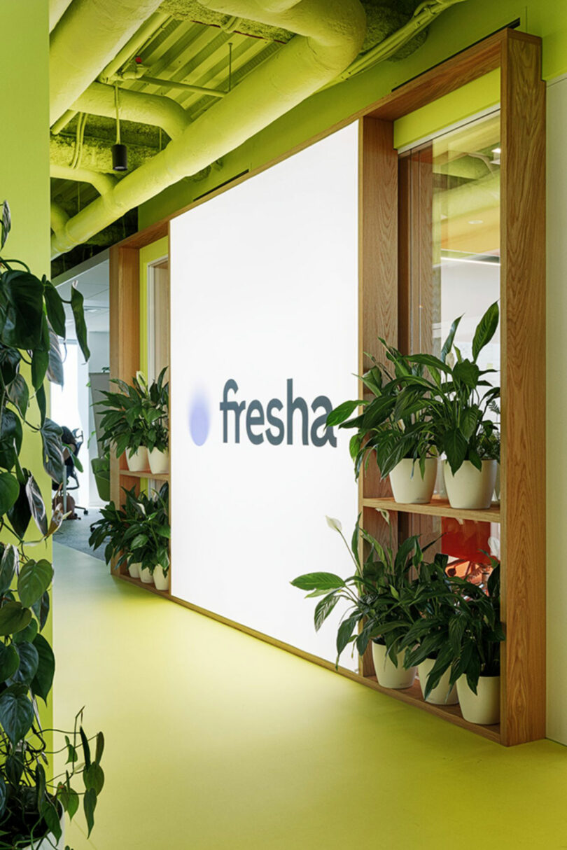 Bright hallway with green walls, plants, and a large illuminated sign displaying the word “fresha.”.