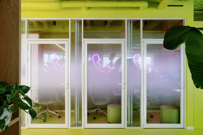 Small office booths with frosted glass doors and neon phone symbols, surrounded by green plants.